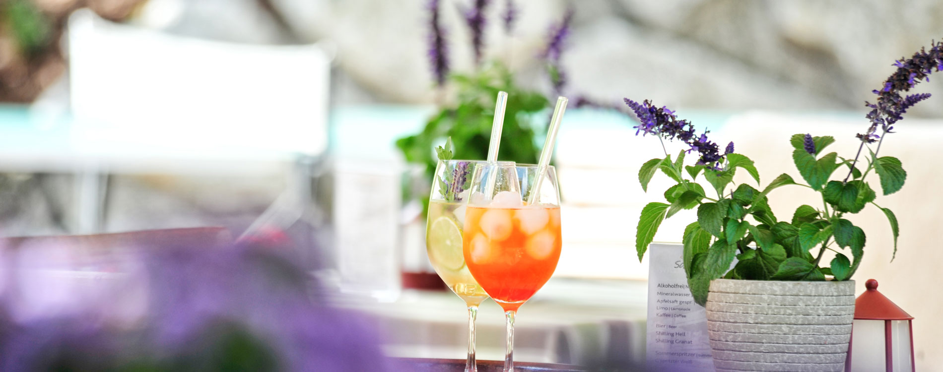 Zwei bunte Sommergetränke mit Eis und Strohhalm auf einem Tisch, umgeben von Kräutern und Lavendel im Blumentopf.