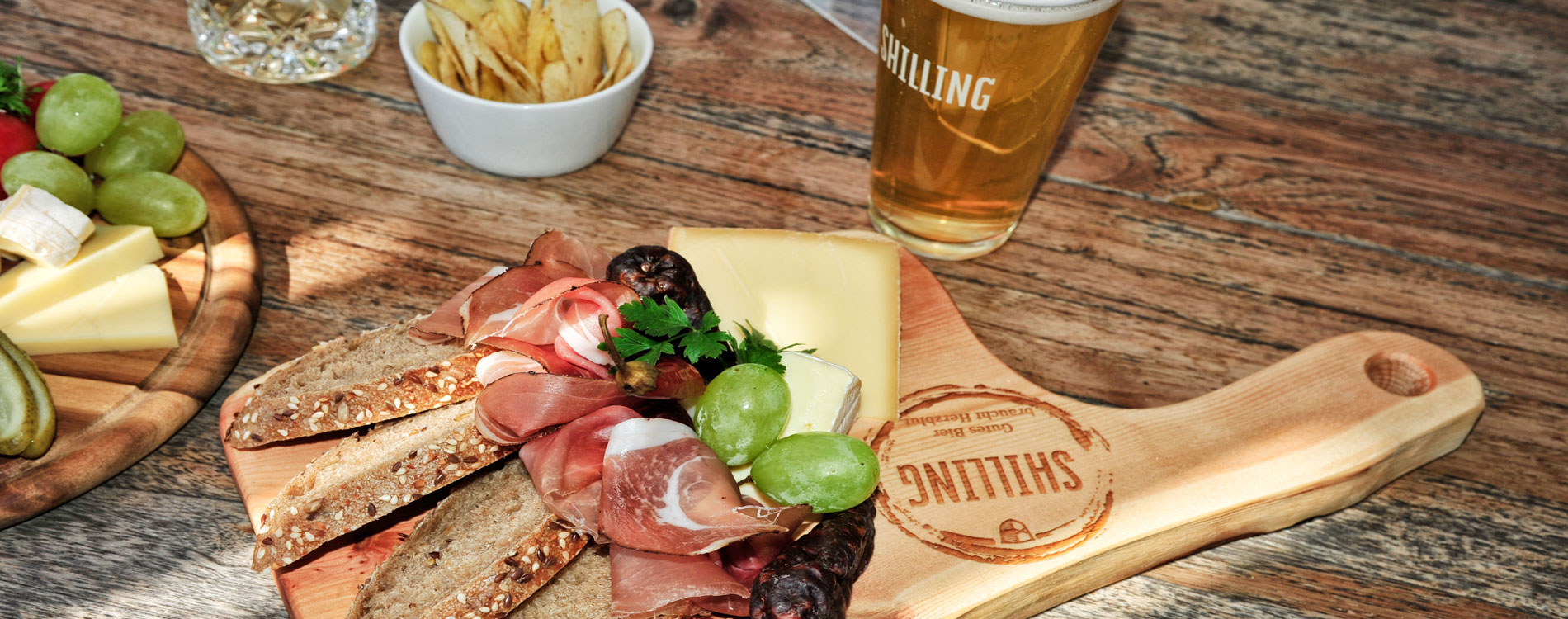 Auf einem Holztisch liegt eine Brettljause mit Brot, Käse, Speck, Trauben und einem Glas Bier mit „SHILLING“-Logo.
