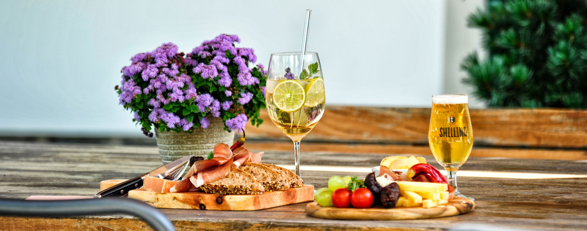 Brotzeit mit Käse, Speck, Obst und Getränken auf rustikalem Holztisch neben Topf mit violetten Blumen.