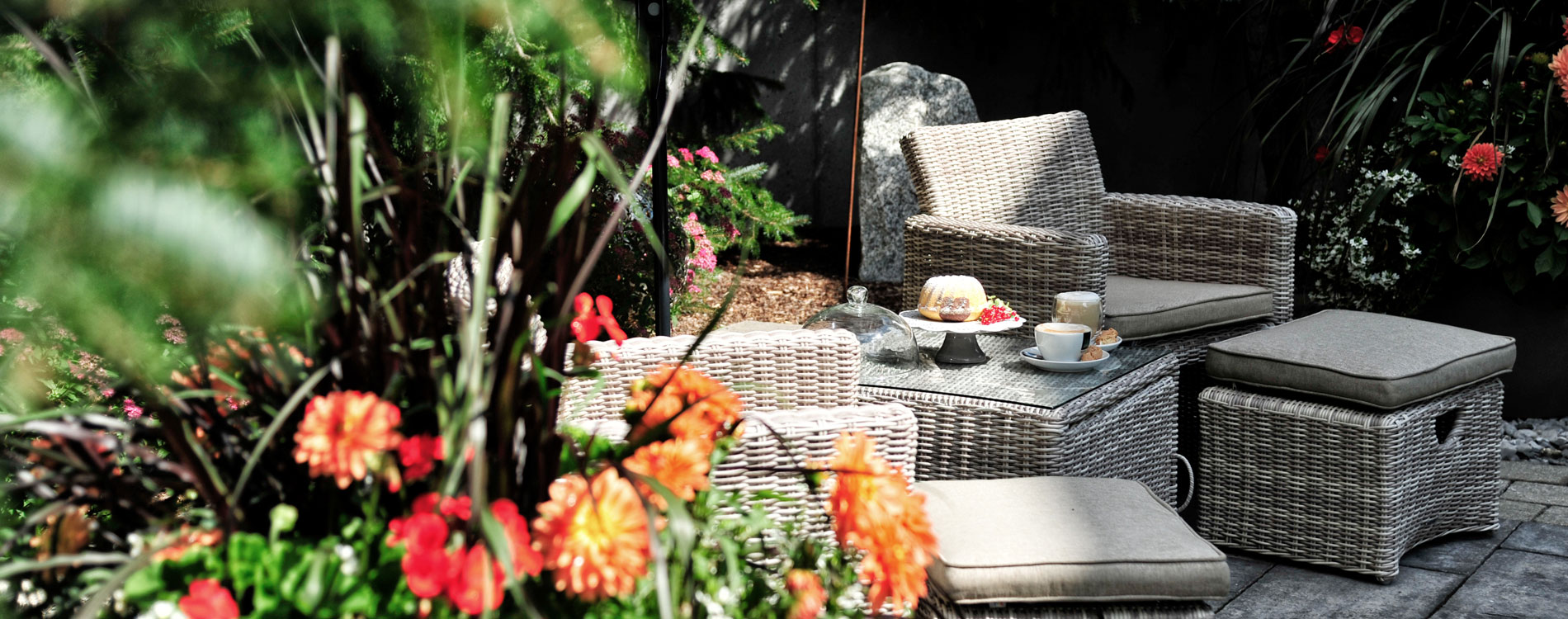 Gemütliche Sitzecke im Garten mit Kaffee, Kuchen und Blumen im Vordergrund, eingerahmt von grünem Pflanzenwuchs.