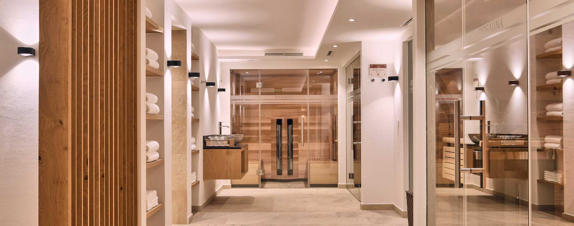 Heller Spa-Bereich mit Holz- und Glaselementen, Blick auf Sauna und Infrarotkabine, Regale mit weißen Handtüchern.
