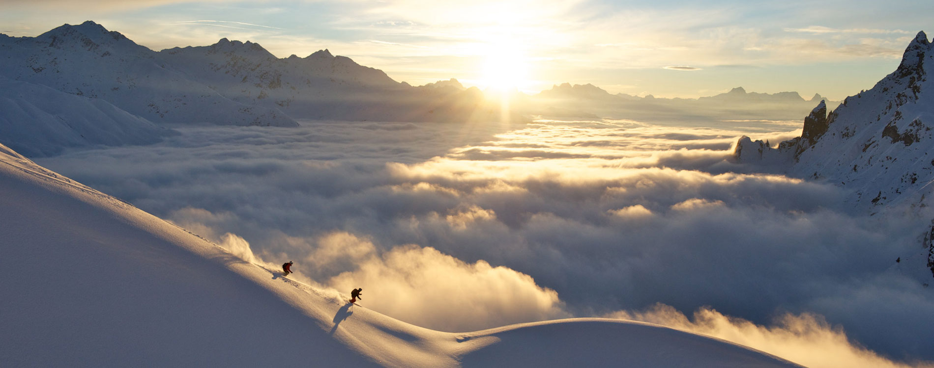 Zwei Skifahrer fahren bei Sonnenaufgang einen verschneiten Hang hinab, unter ihnen ein Meer aus Wolken in den Bergen.
