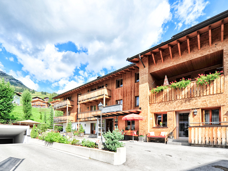 Außenansicht des Chalets Anna Maria in Lech mit Balkonen, roten Sonnenschirmen und blühenden Blumen bei blauem Himmel.