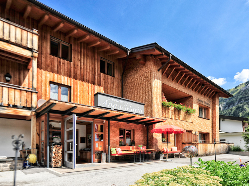 Eingangsbereich des Apartmenthauses Anna Maria mit Holzfassade, Sitzbank und Sonnenschirm bei strahlendem Wetter.
